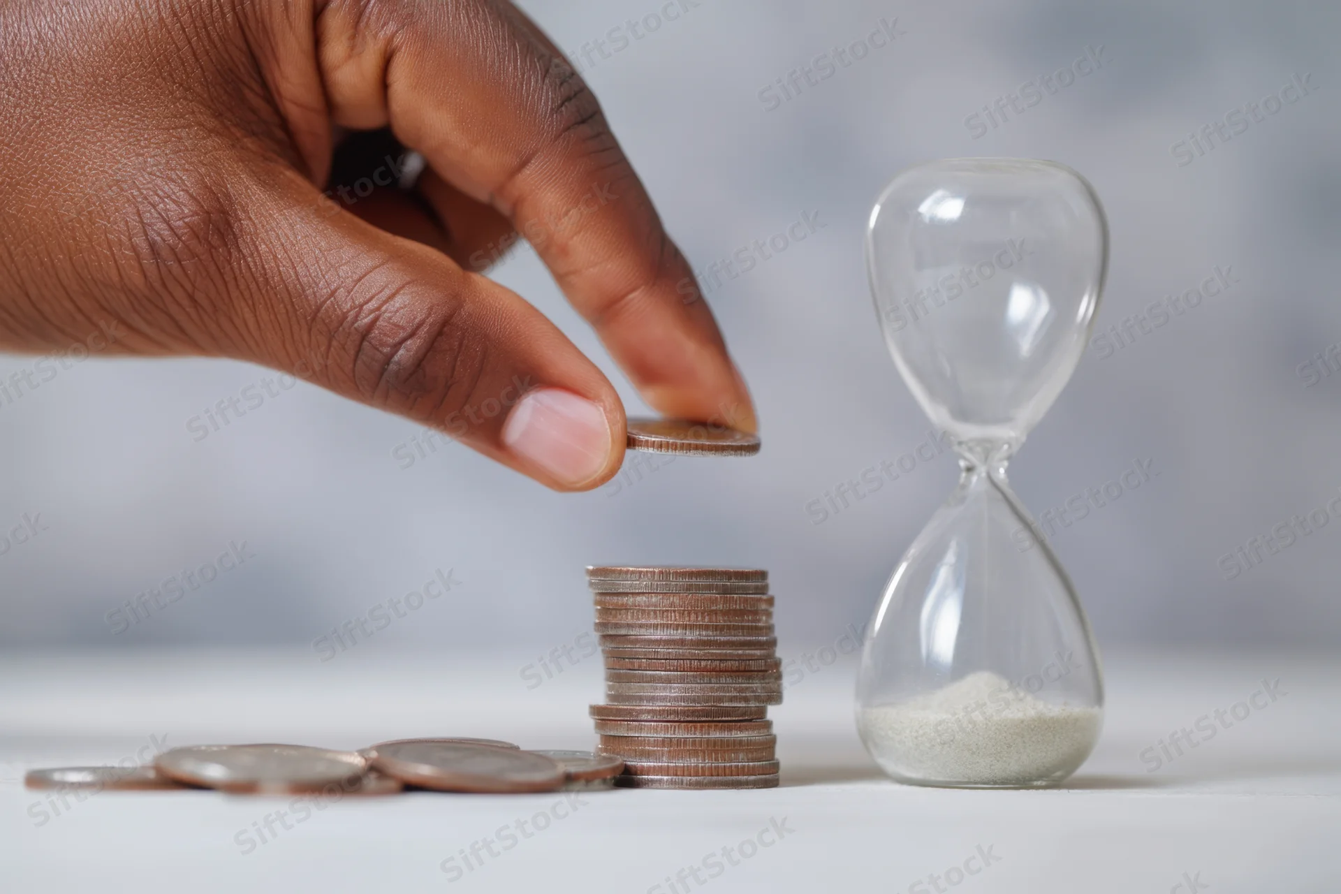 Hand placing coin on growing stack beside hourglass on white surface