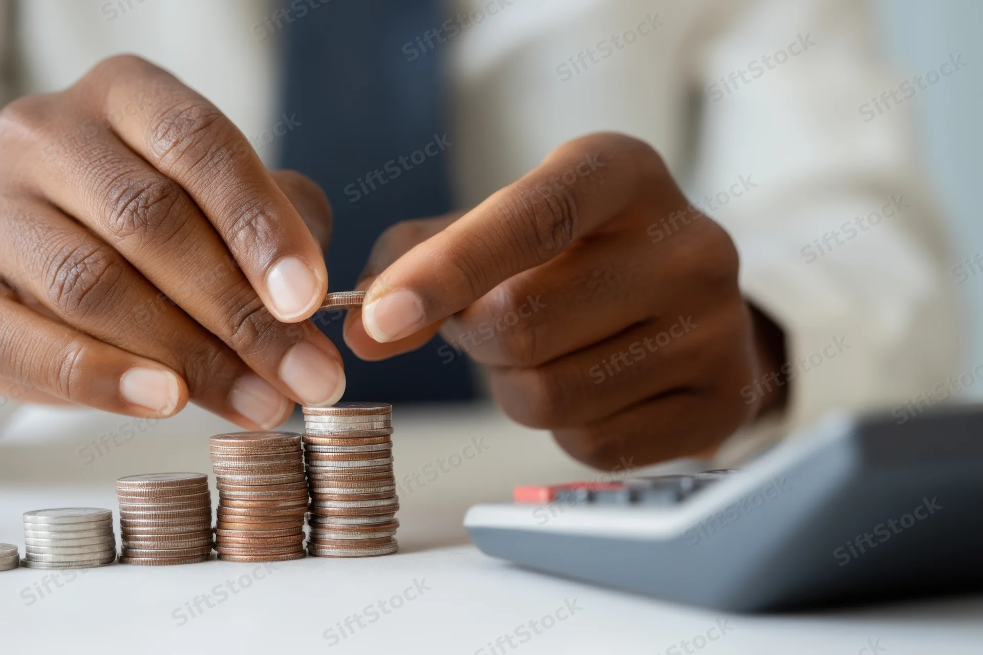 Hands stacking coins in increasing height on a desk beside a calculator