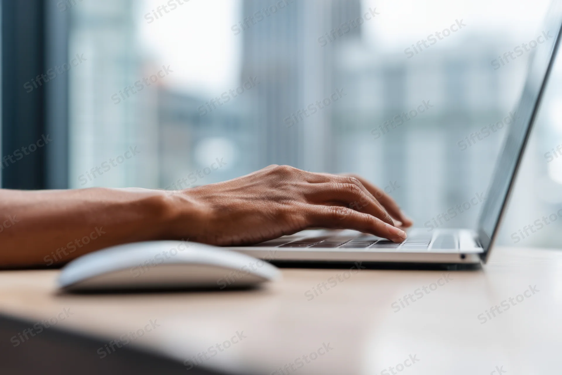 Hands typing on a laptop keyboard at a desk near a window with city view