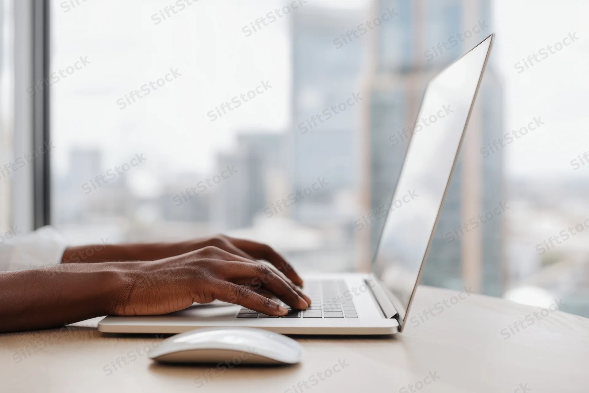 Hands typing on a sleek silver laptop at a desk with city view through window