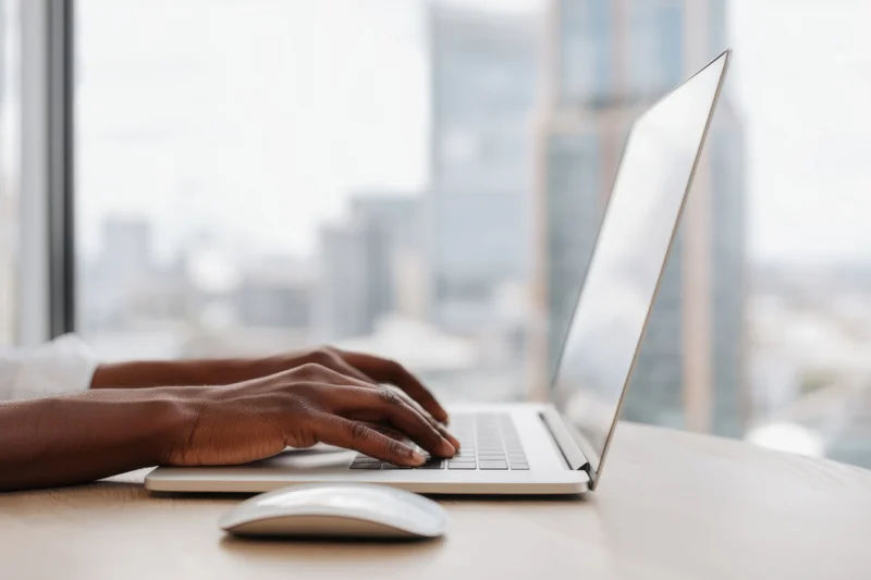 Hands typing on a sleek silver laptop at a desk with city view through window