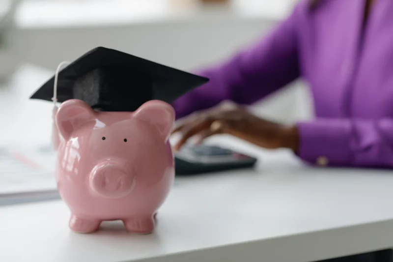 Pink piggy bank wearing graduation cap on desk with woman using calculator in office