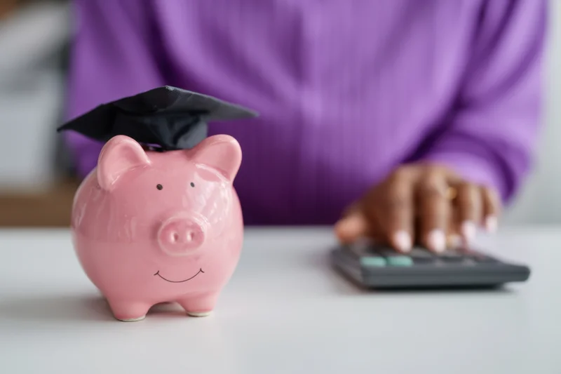 Pink piggy bank wearing graduation cap on desk with hand using calculator nearby