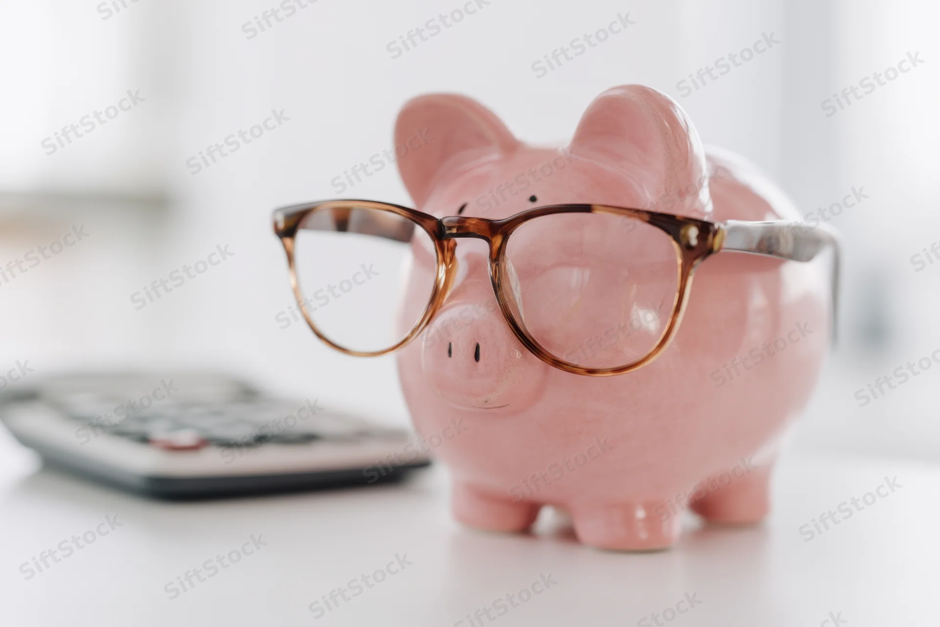 Pink piggy bank wearing tortoiseshell glasses beside a calculator on a desk