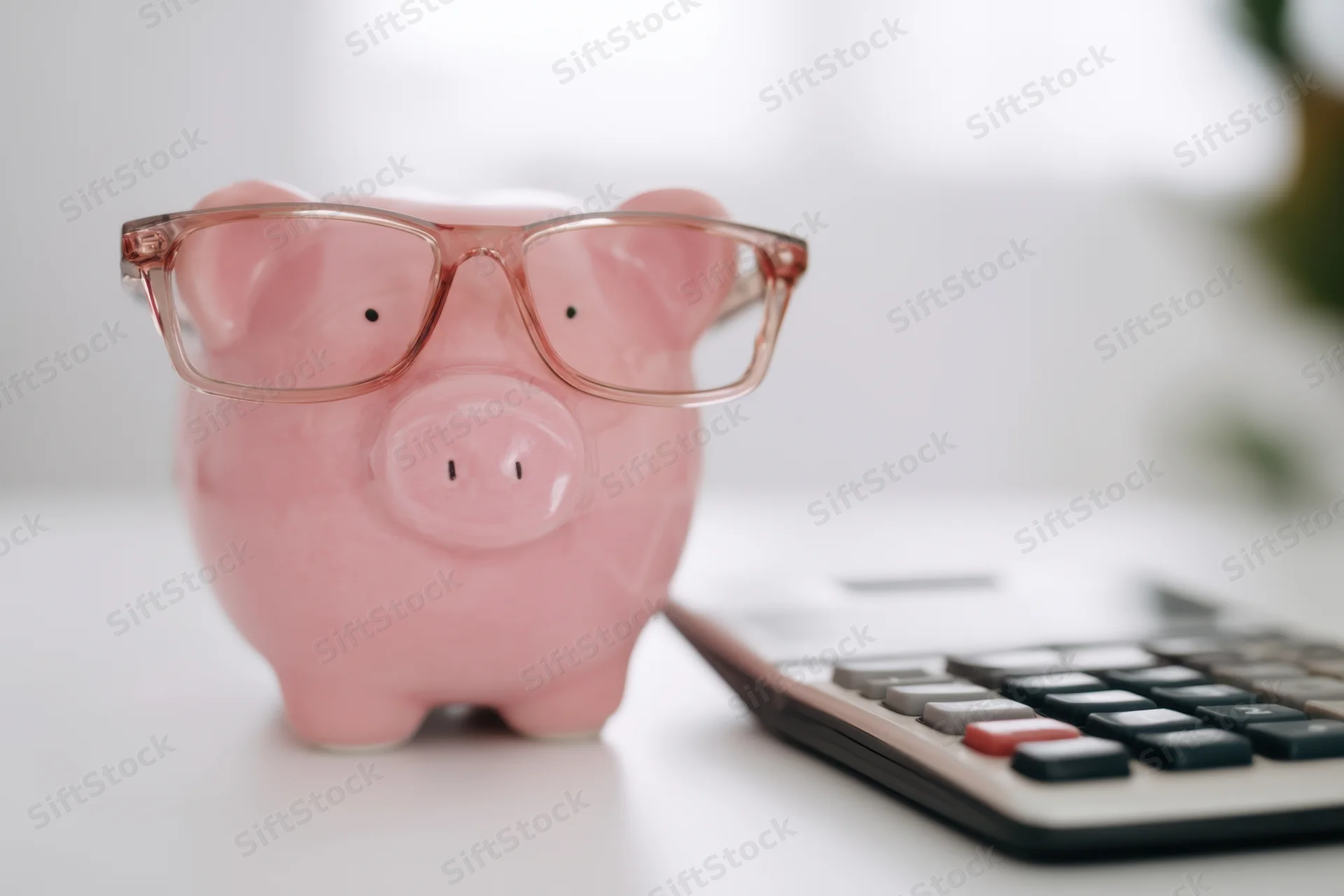 Pink piggy bank wearing glasses placed next to a calculator on a white surface
