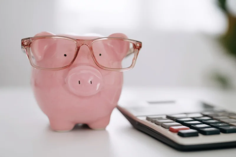 Pink piggy bank wearing glasses placed next to a calculator on a white surface