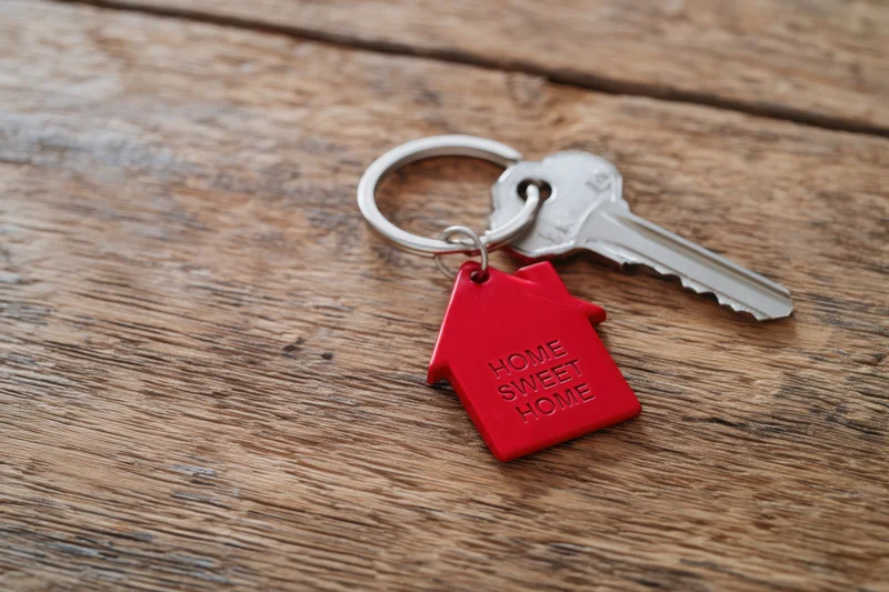 Red house-shaped keychain with home sweet home inscription attached to a key resting on a wooden surface