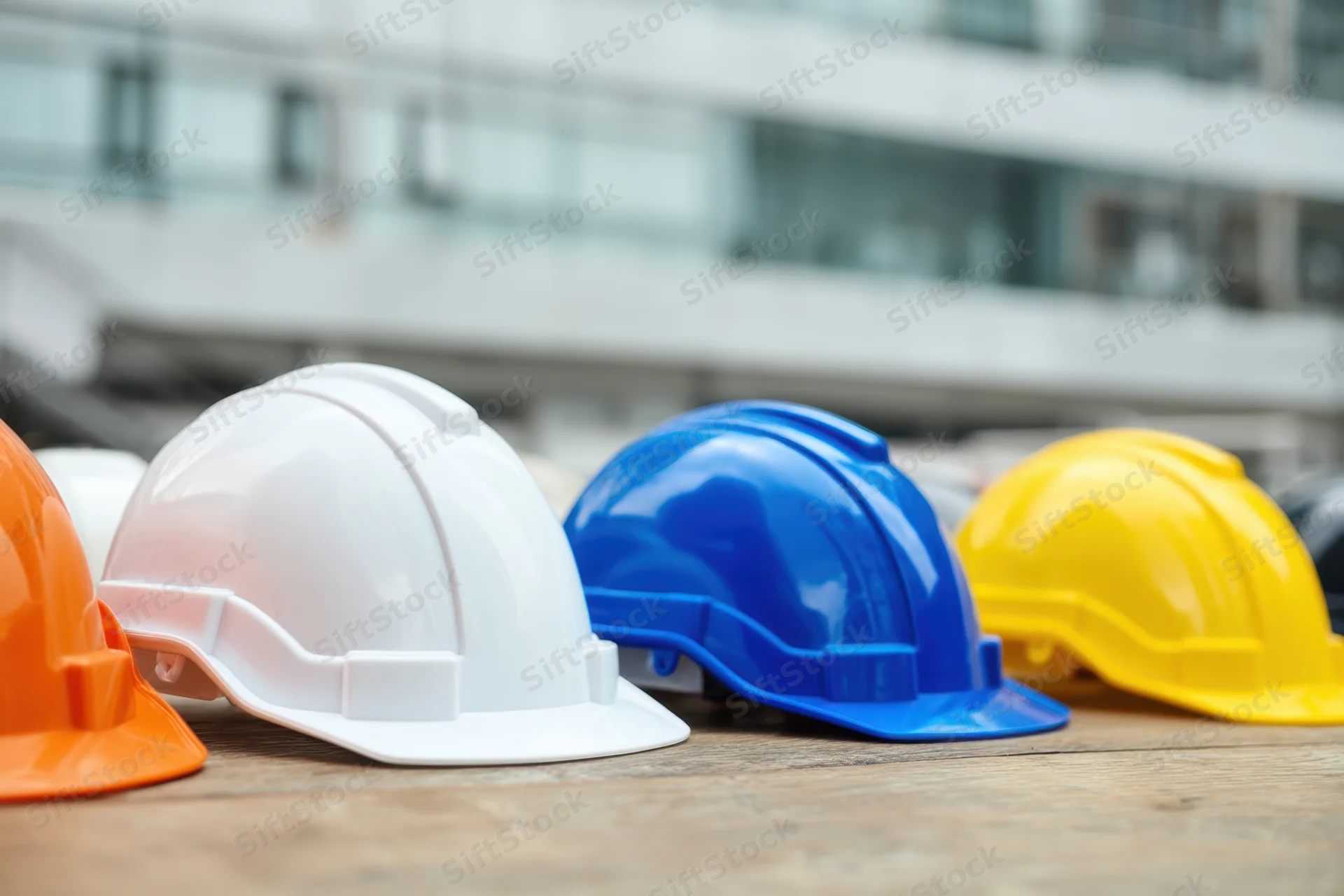 Row of colorful construction hardhats arranged on wooden surface with blurred building in background