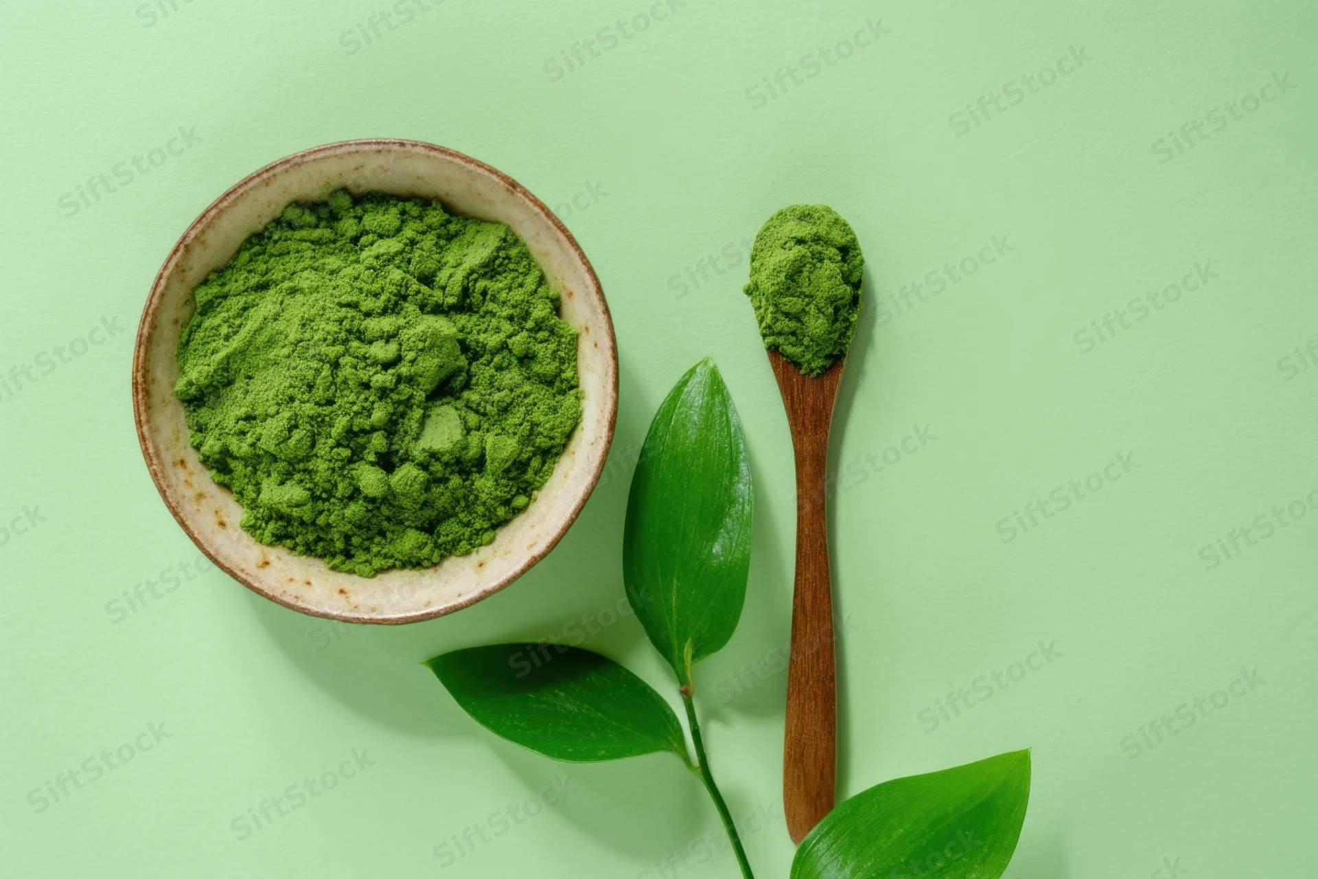 Matcha powder in a ceramic bowl with wooden spoon and green leaves on a light green surface