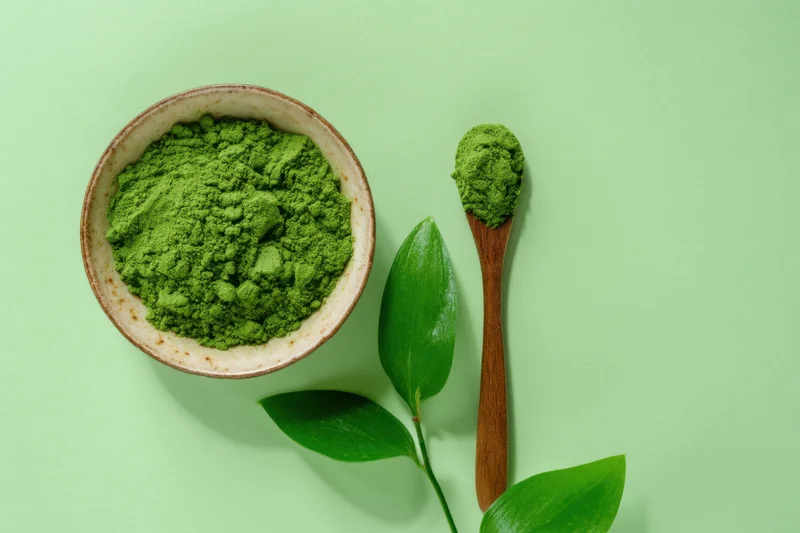 Matcha powder in a ceramic bowl with wooden spoon and green leaves on a light green surface