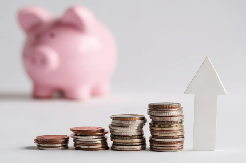 Stacks of coins arranged in ascending order with a white arrow and a pink piggy bank in the background