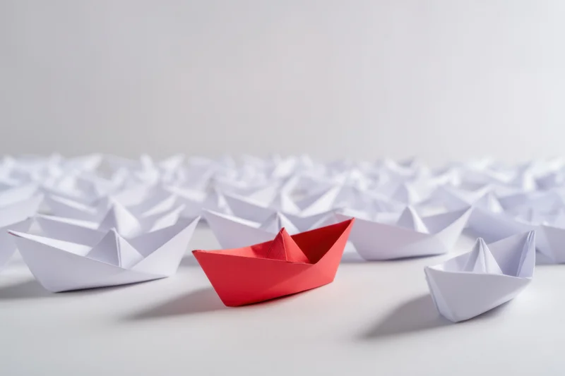 Red paper boat among white boats on a plain surface