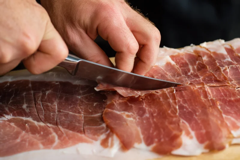Hand slicing cured ham on a wooden board with a sharp knife