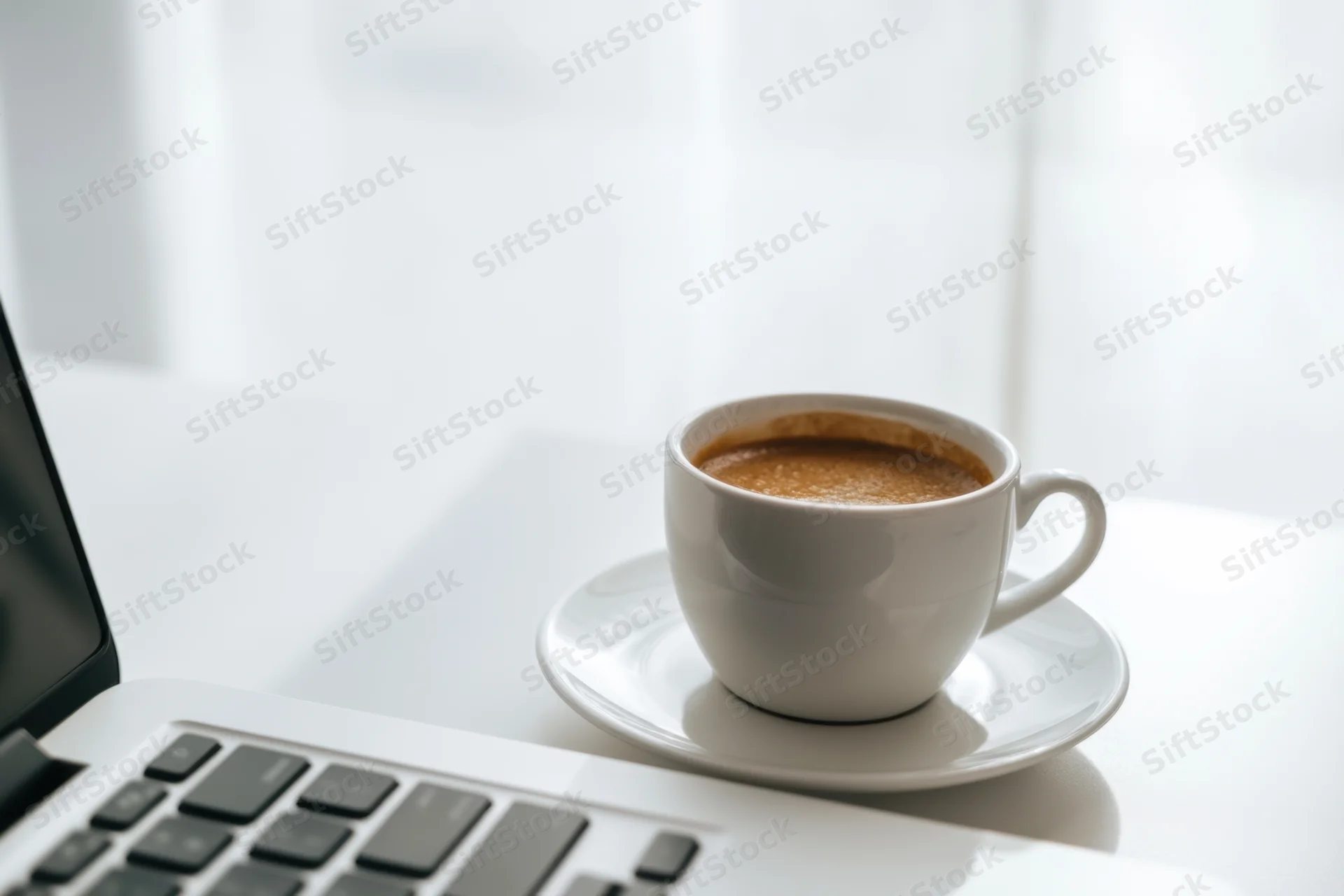 Laptop keyboard beside a white coffee cup on a bright desk