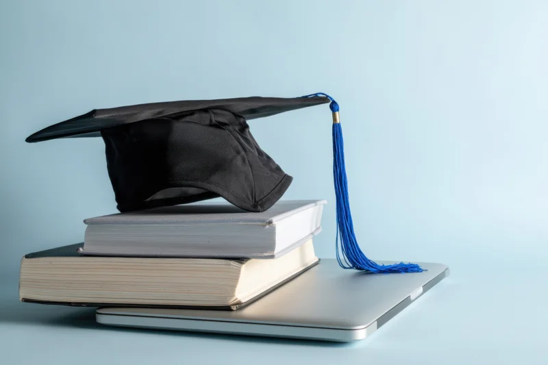 Graduation cap with blue tassel resting on stacked textbooks atop a closed silver laptop on a light blue surface