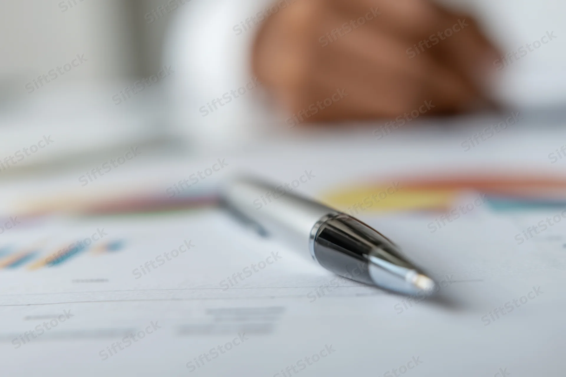 Silver pen resting on financial report with charts and graphs on desk, shallow focus, hand blurred in background