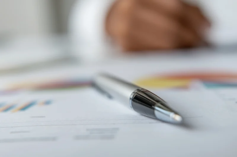 Silver pen resting on financial report with charts and graphs on desk, shallow focus, hand blurred in background