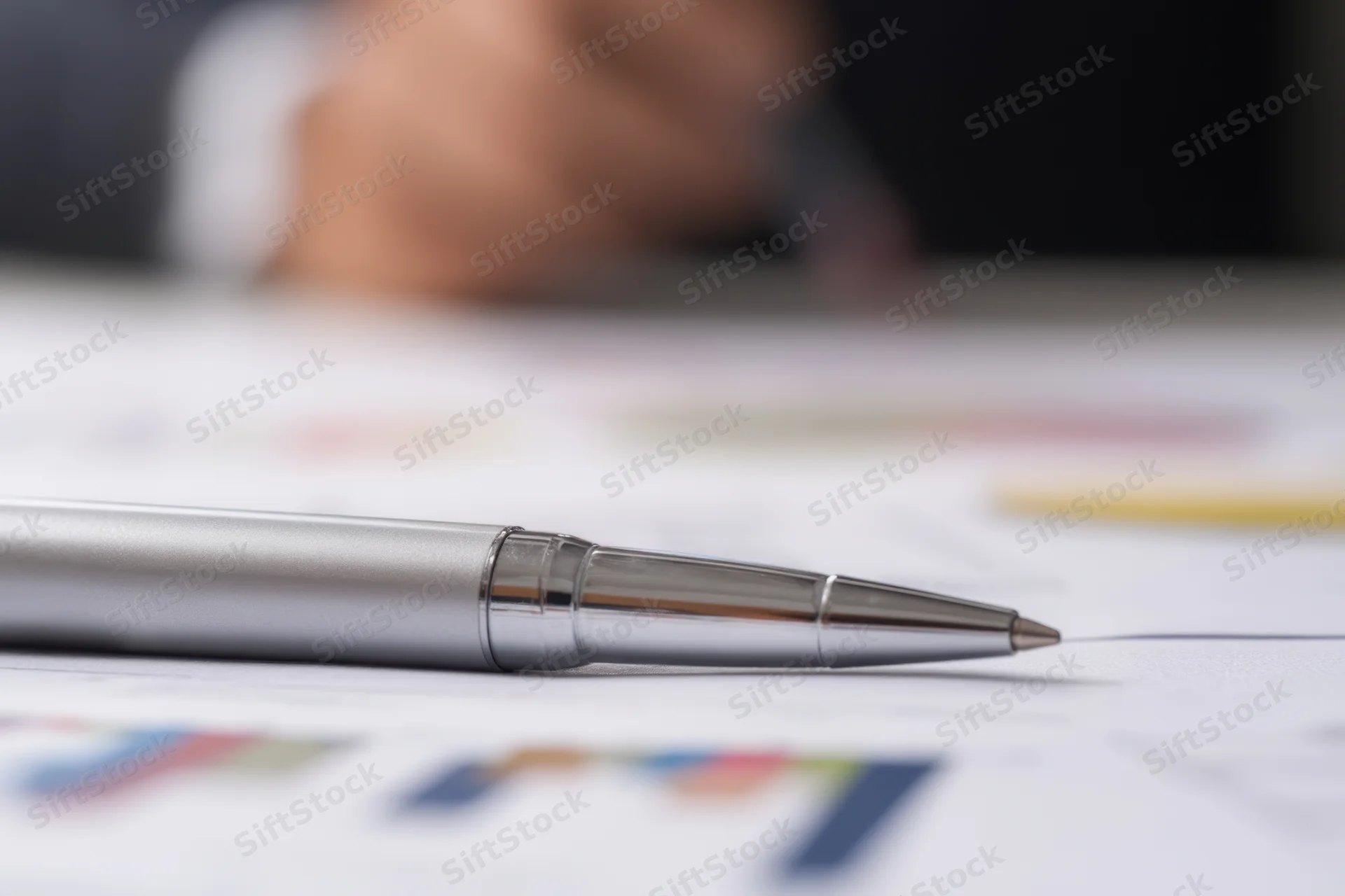 Silver pen resting on financial report with charts and graphs on desk, hand blurred in background