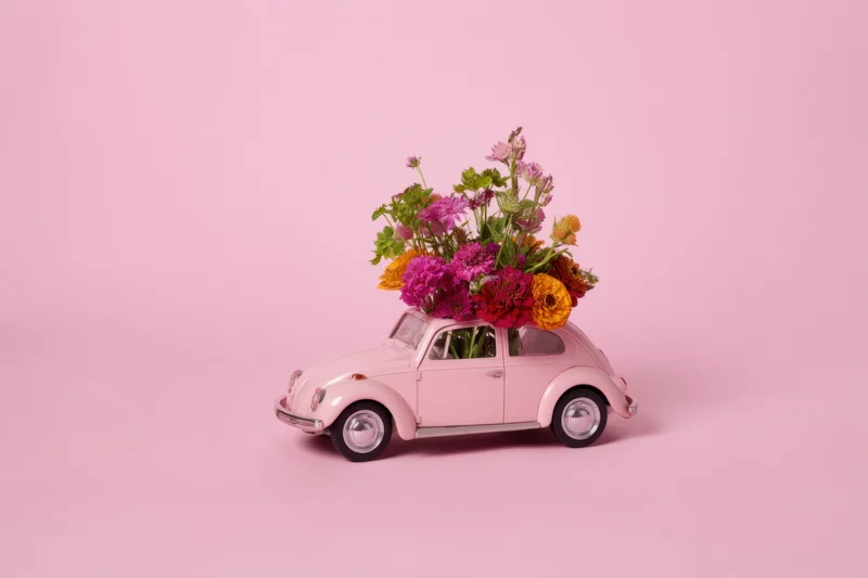Pastel pink toy beetle car carrying a vibrant floral bouquet on its roof against a plain pink background