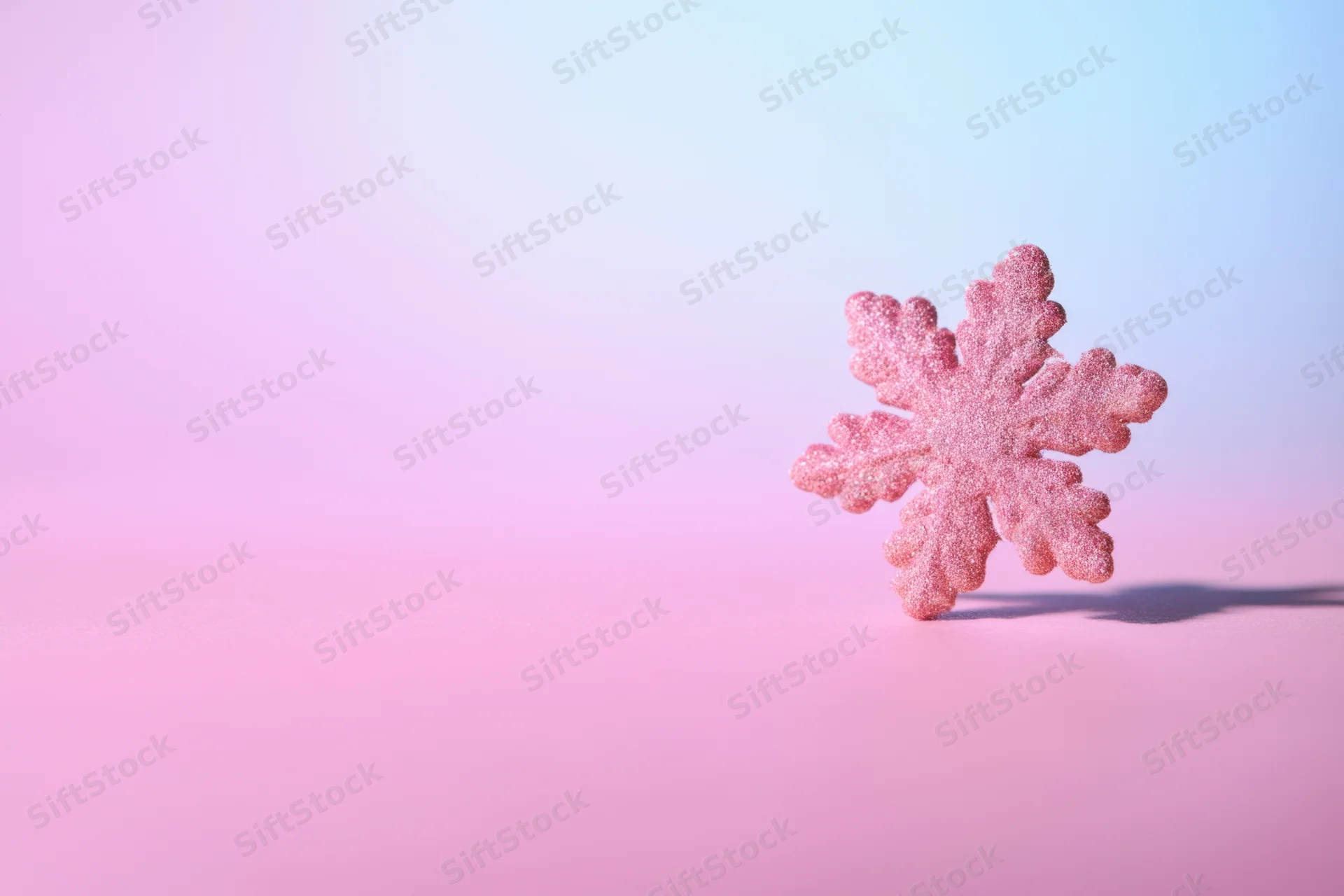 A sparkling pink snowflake ornament standing on a soft pink to blue gradient background with copyspace