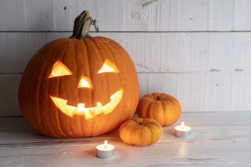 Carved pumpkin with glowing face and candles on white wooden surface