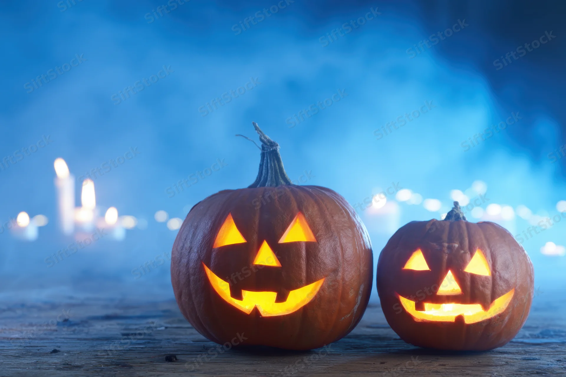 Two carved pumpkins glowing with candlelight on a wooden surface in a smoky blue setting