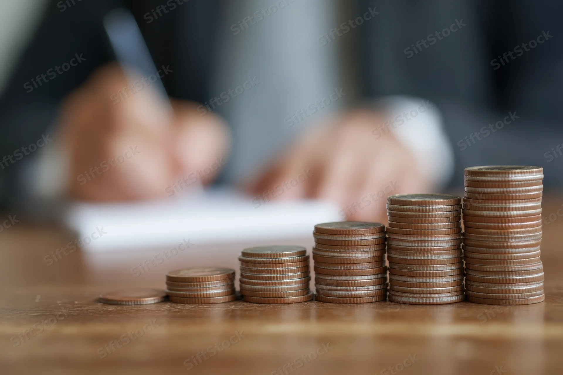 Stacks of coins arranged in ascending order on a wooden table with a person writing in the background