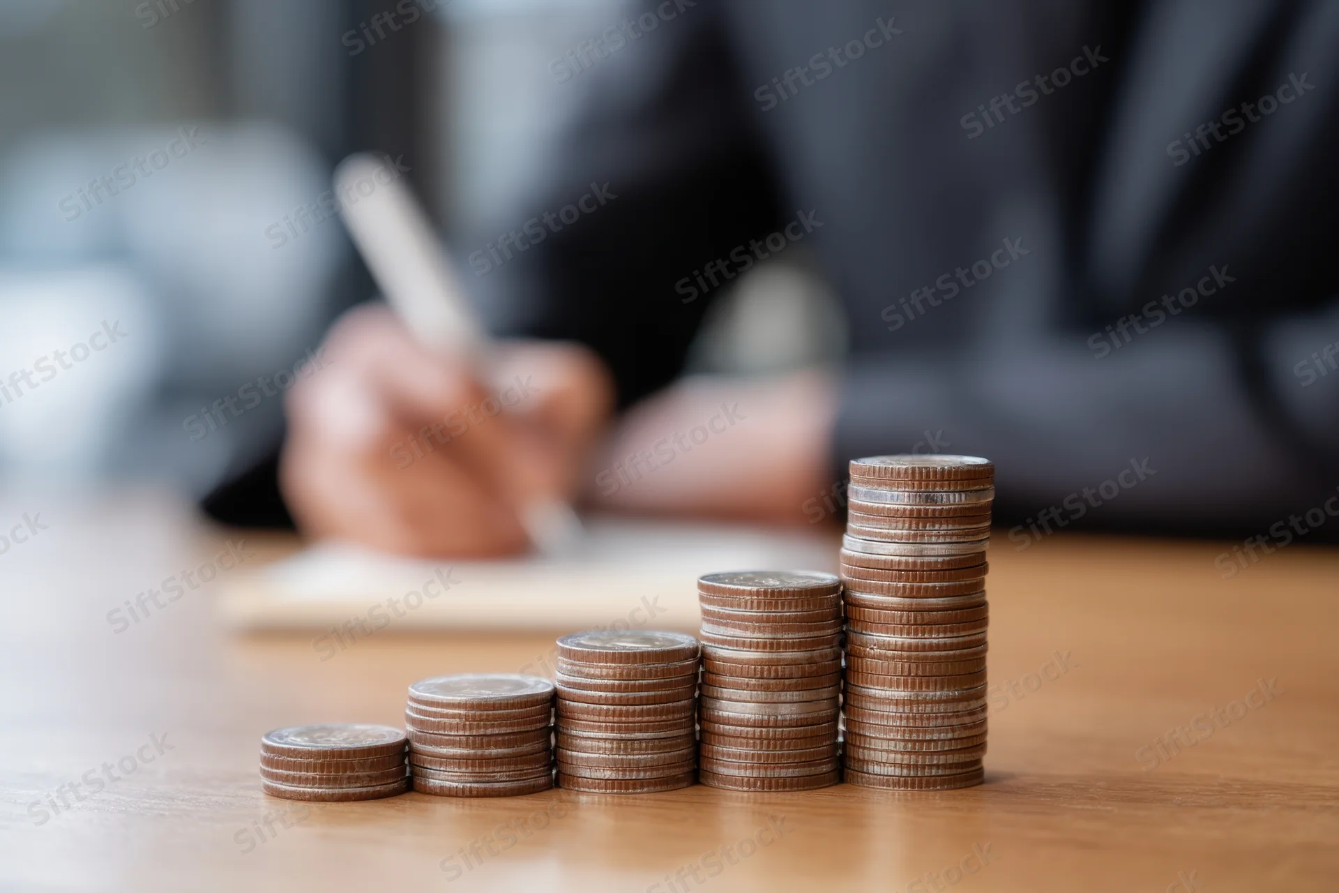 Stacks of coins arranged in ascending order on a wooden table with a person in a suit writing in the background
