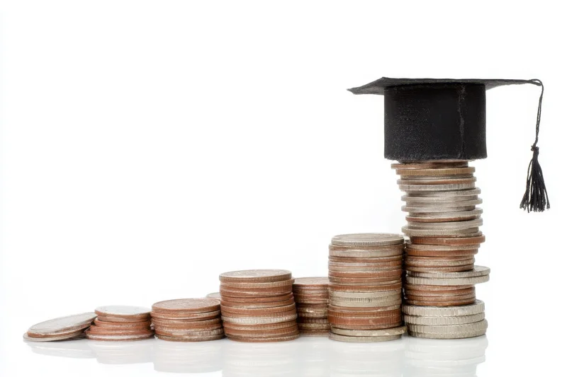 Stacks of coins arranged in ascending order topped with a graduation cap on a white surface