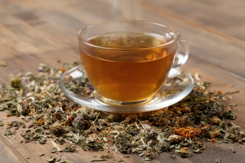 Steaming cup of herbal tea on glass saucer surrounded by dried herbs on wooden table
