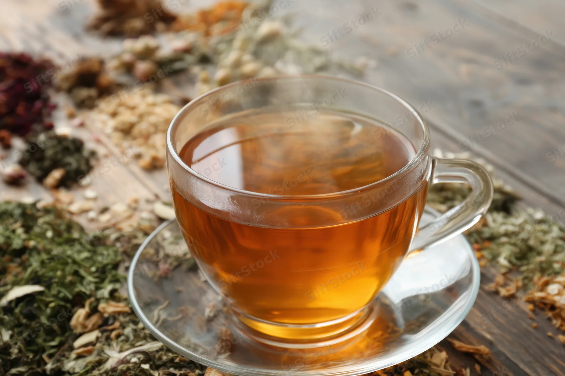 Steaming cup of herbal tea on glass saucer surrounded by dried herbs on wooden table