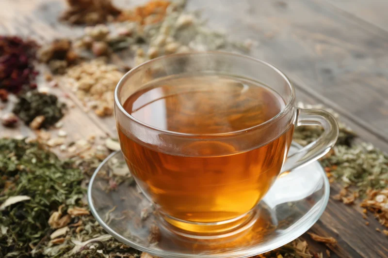 Steaming cup of herbal tea on glass saucer surrounded by dried herbs on wooden table
