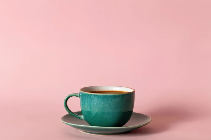 Teal ceramic coffee cup on matching saucer against pink background