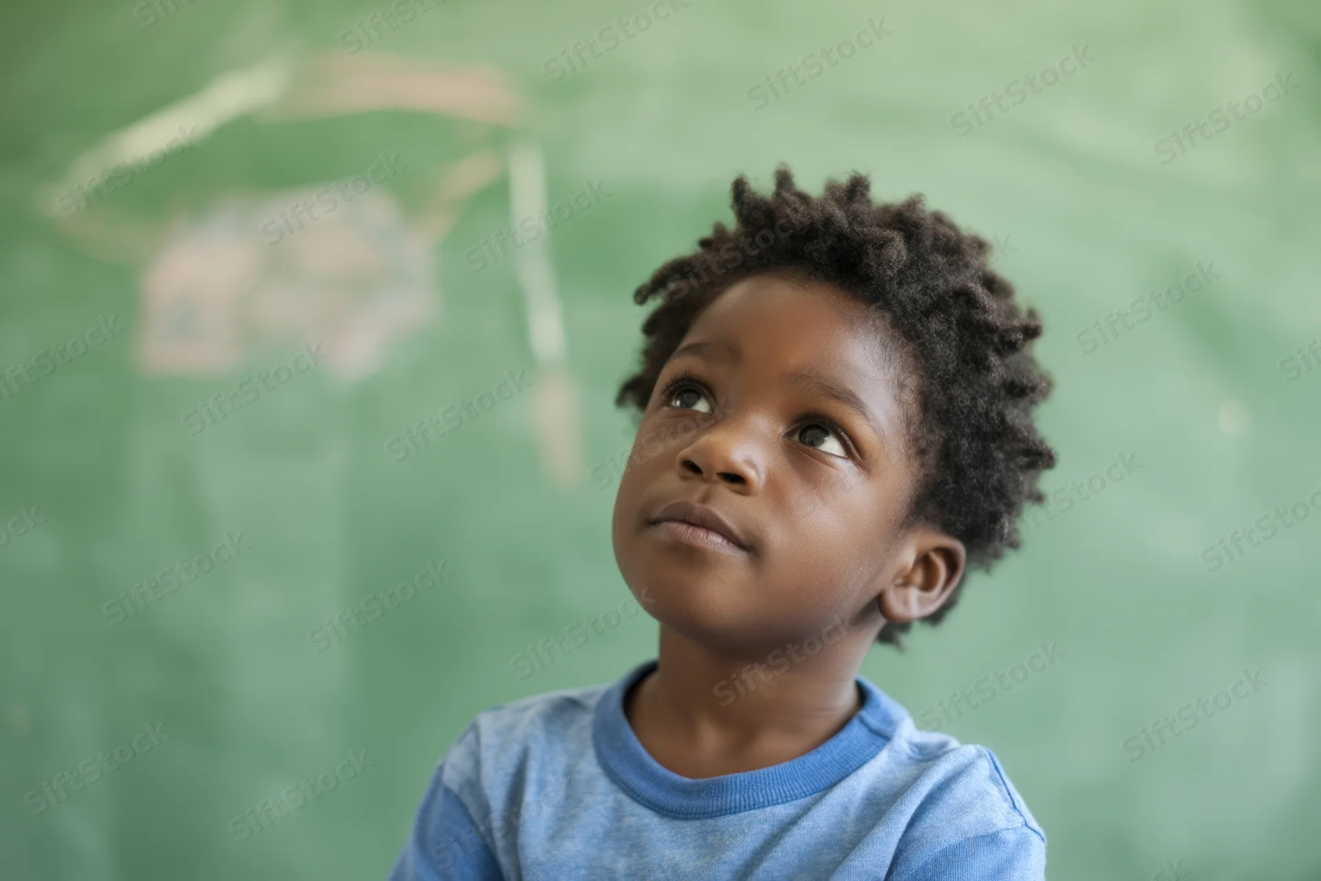 Young black boy with thoughtful expression looking up in classroom near green chalkboard