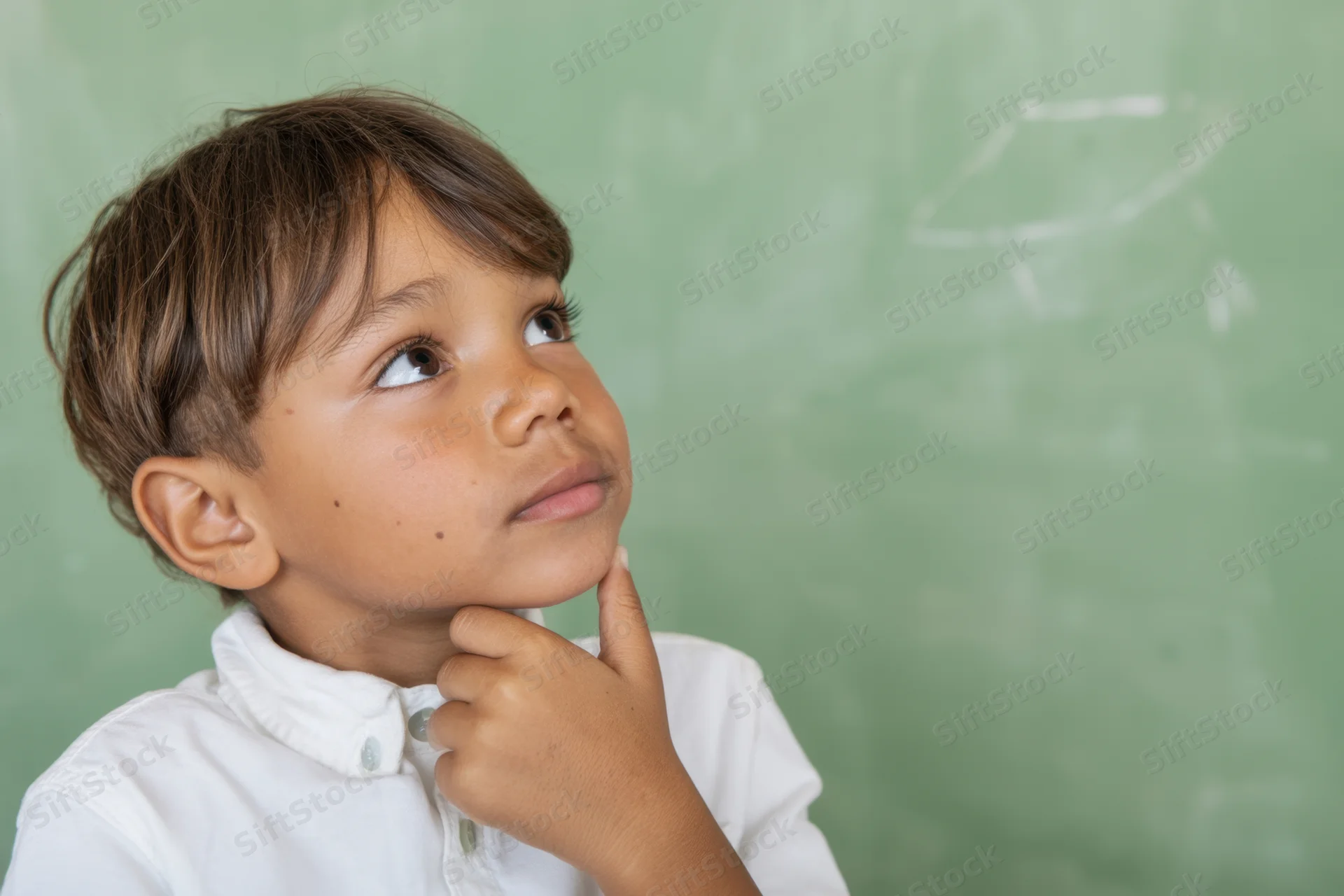 Young boy with hand on chin looking thoughtfully at chalkboard in classroom