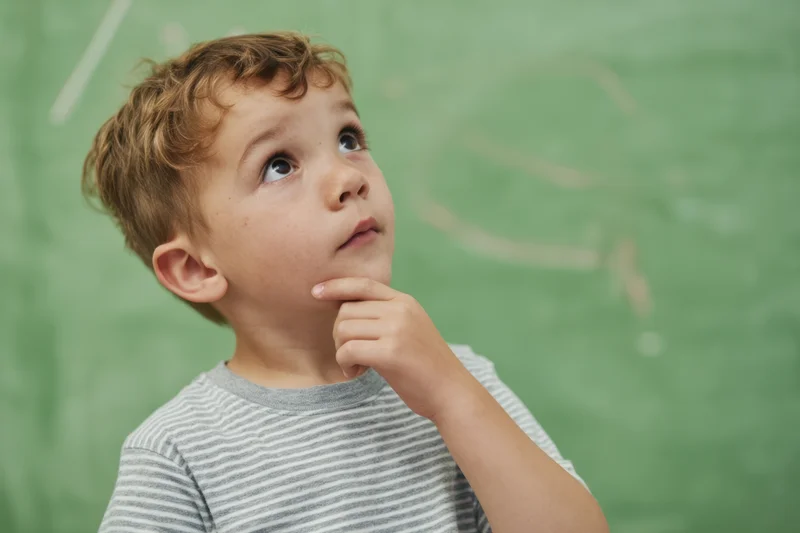 Young boy with hand on chin looking upward in classroom setting