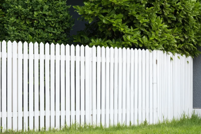 White picket fence along a grassy yard with green shrubs behind it