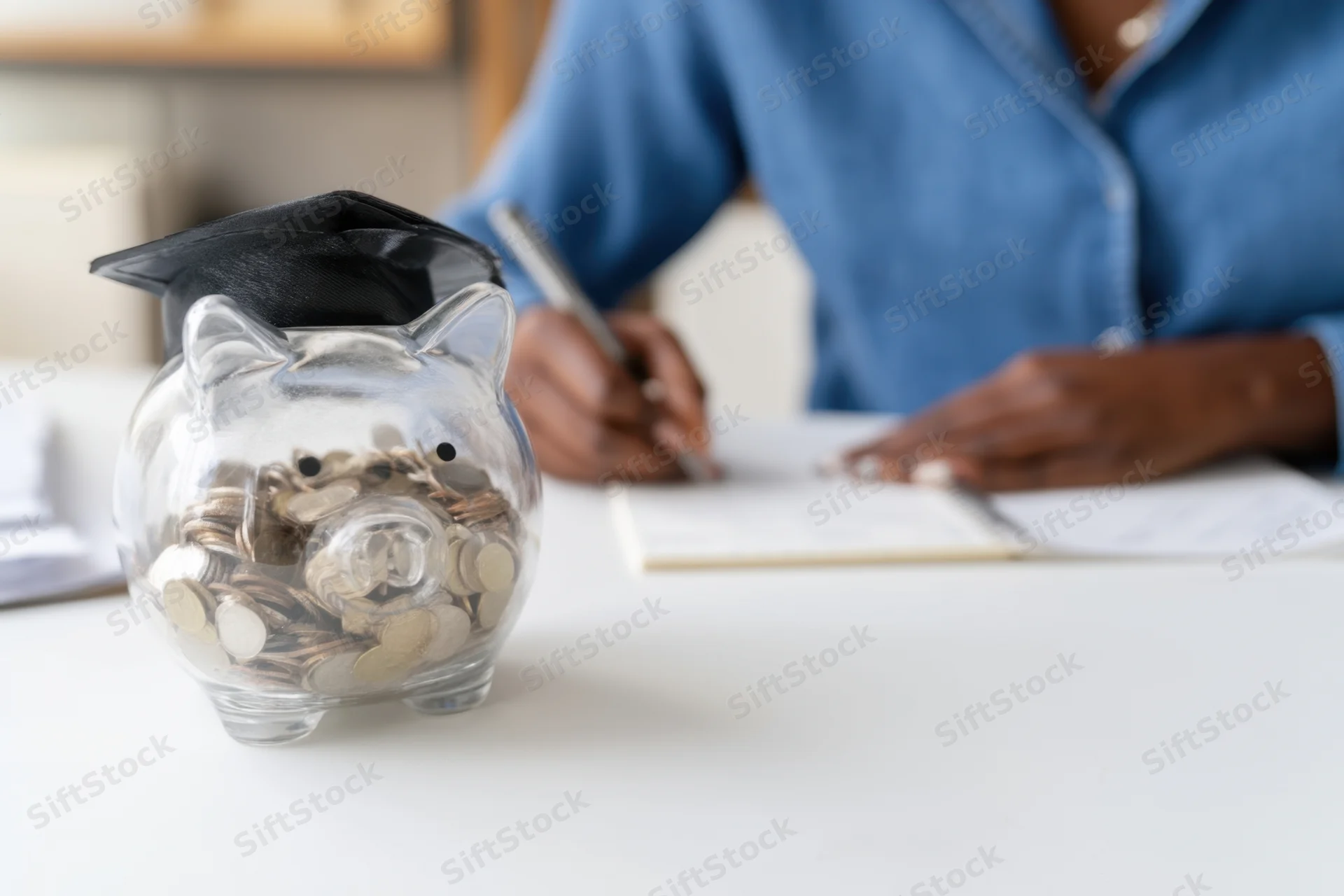 Transparent piggy bank with graduation cap filled with coins on desk beside woman writing in notebook