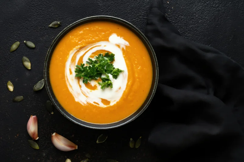 Orange soup with cream swirl and parsley garnish in a bowl on dark surface with scattered seeds and garlic cloves