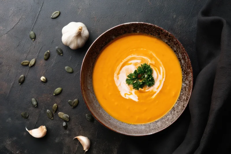 Orange pumpkin soup in a ceramic bowl topped with cream swirl and parsley garnish on a dark textured surface with scattered garlic and seeds
