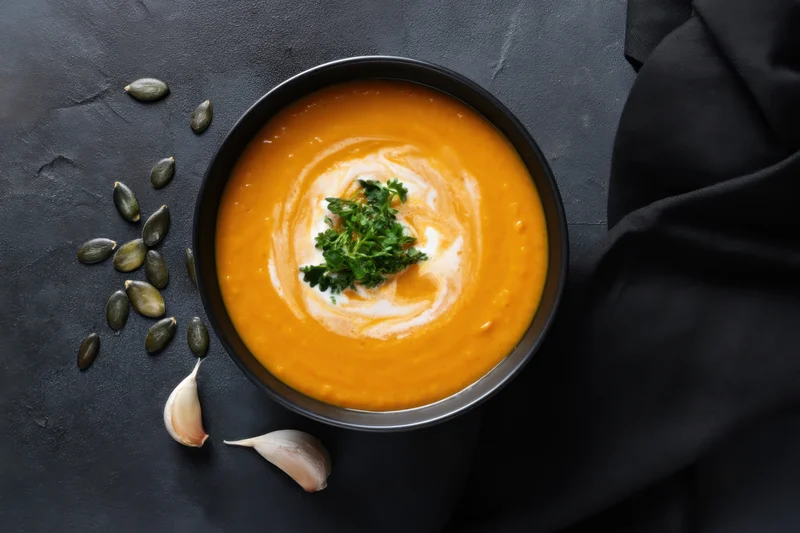 Bowl of orange pumpkin soup topped with cream swirl and parsley garnish on dark surface with garlic and seeds nearby