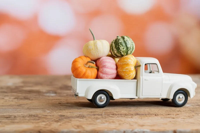Vintage white toy truck carrying small colorful pumpkins and gourds on a wooden surface with blurred autumn background