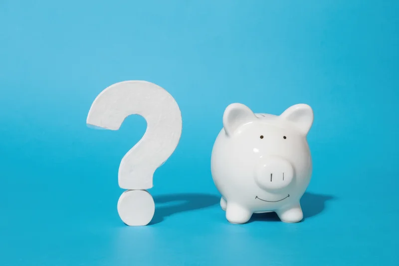 White ceramic piggy bank with friendly expression beside a large white question mark on a solid blue background