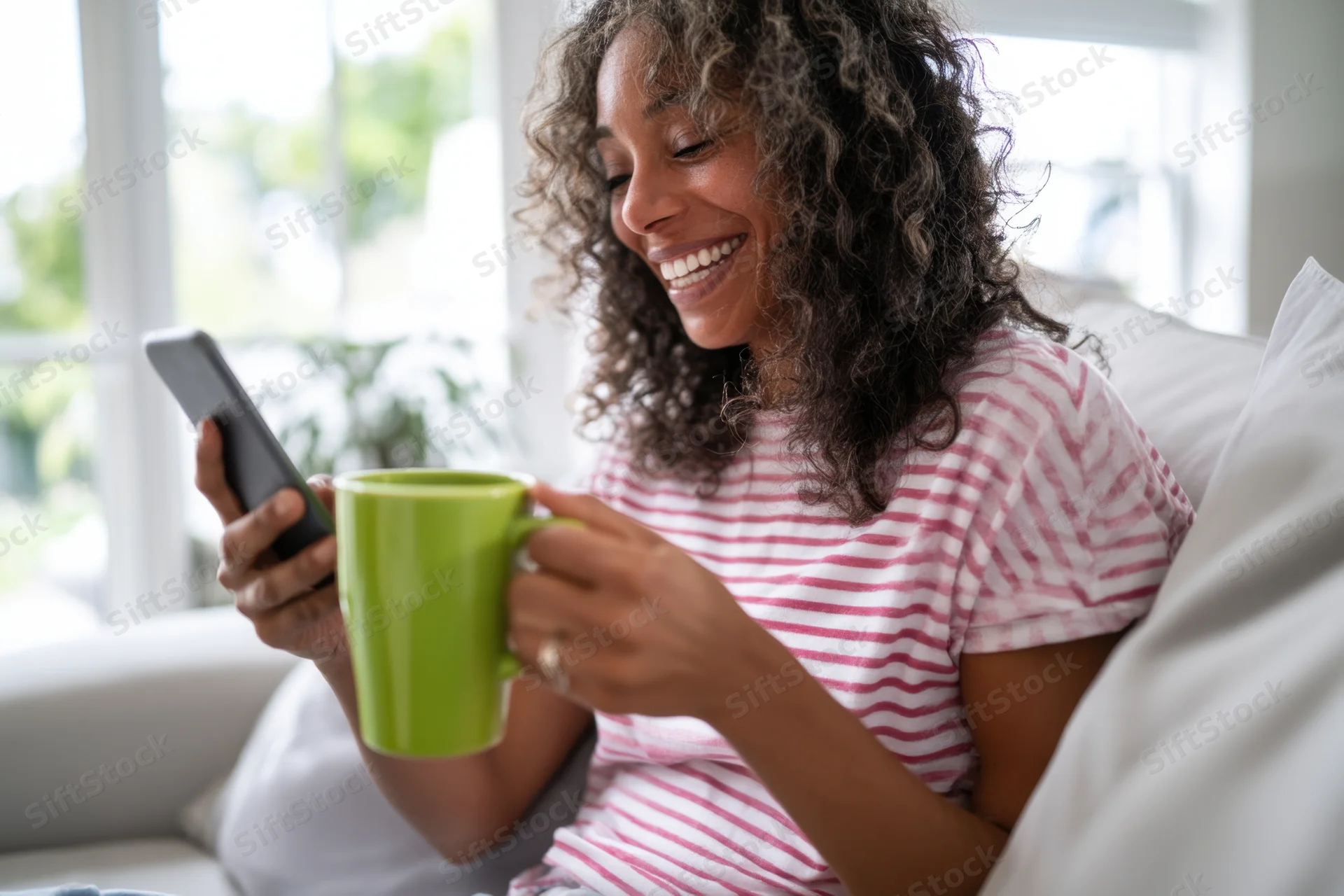 Woman smiling while holding smartphone and green mug on sofa at home