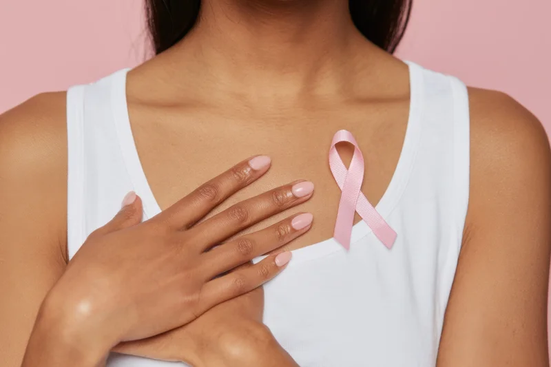Woman in white tank top touching her chest with pink ribbon pinned nearby