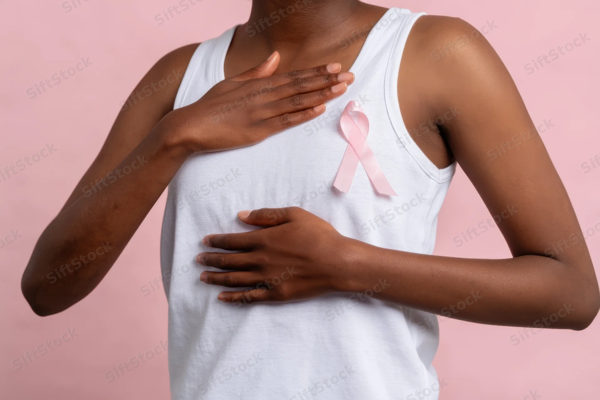 Woman in white tank top touching her chest with pink ribbon pinned to her shirt