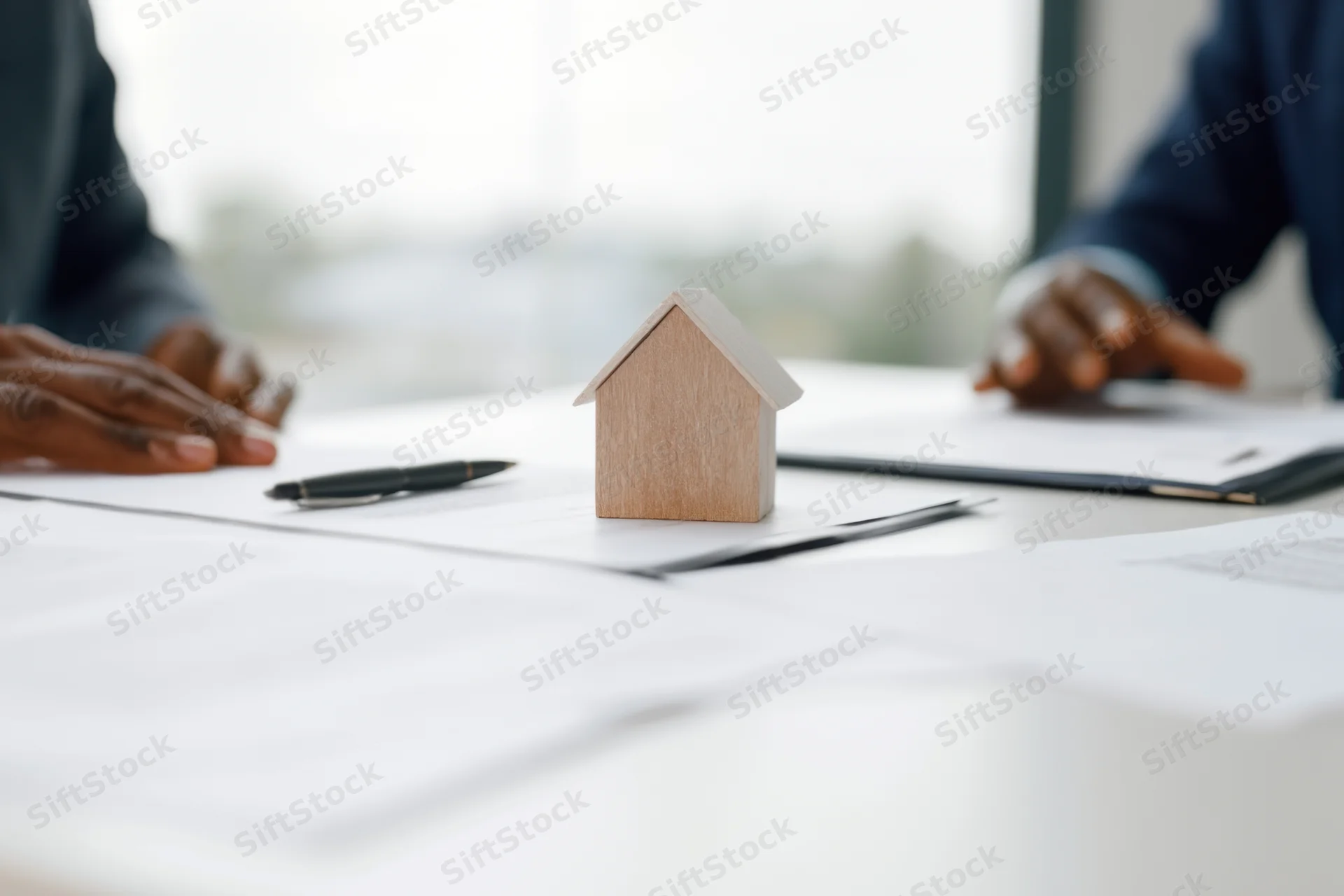 Wooden house model on desk surrounded by documents and pens during a real estate meeting