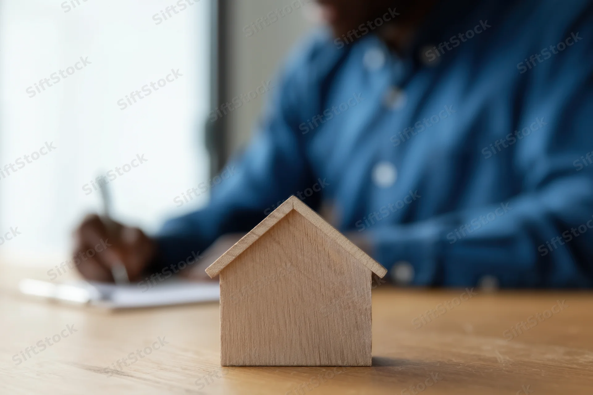 Wooden miniature house on desk with man writing in notebook in background