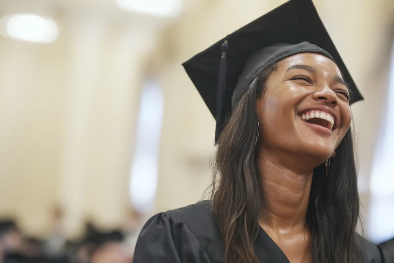 Young black woman in graduation cap and gown laughing joyfully at ceremony