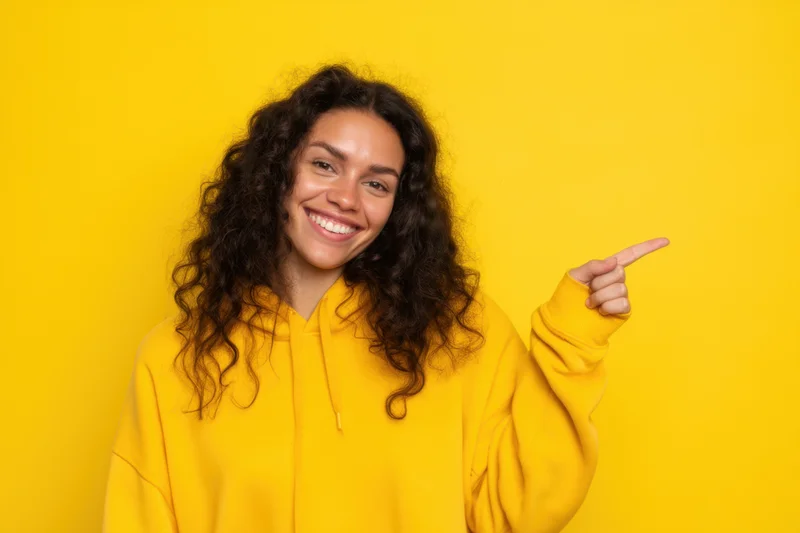 Young woman in yellow hoodie smiling and pointing to side against yellow background